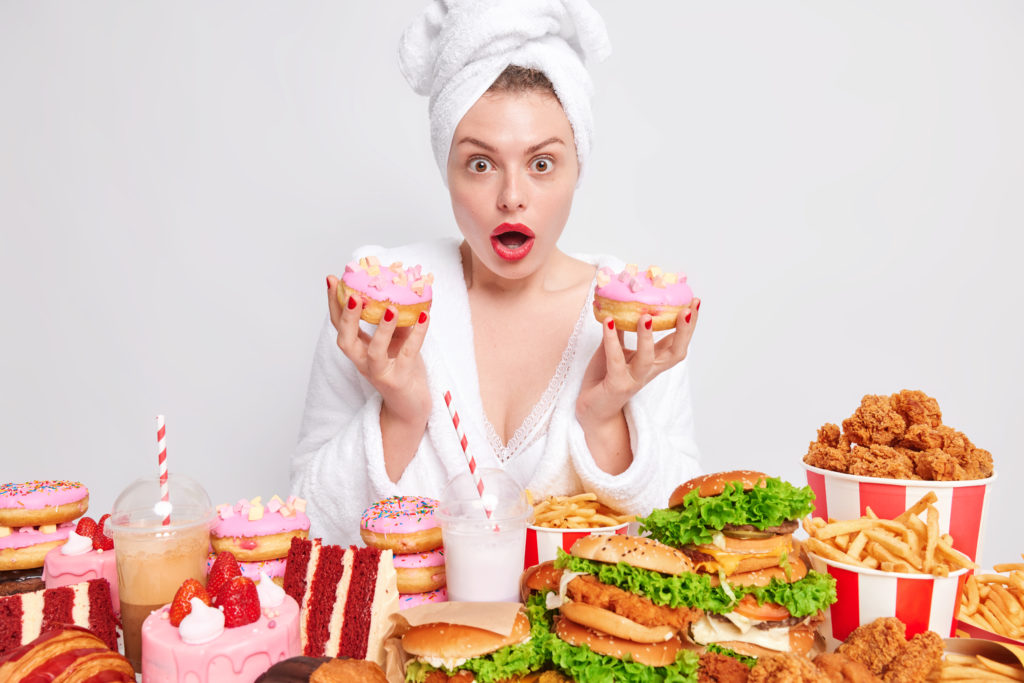 Shocked hungry woman eats high-calorie fatty food holds two doughnuts looks wondered expression