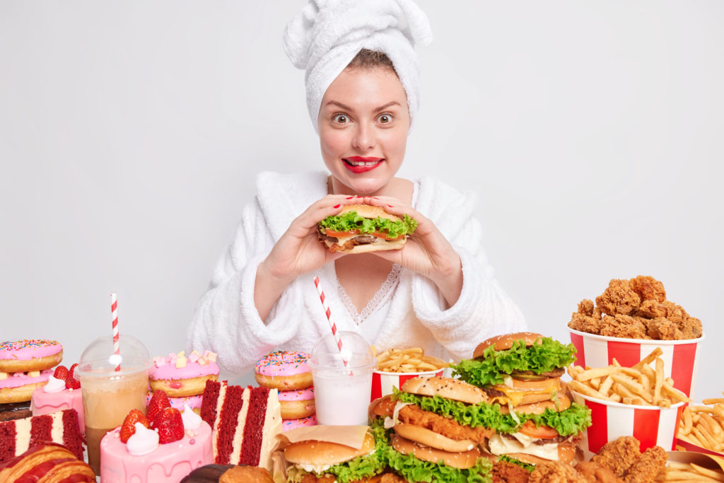 woman eats greedily burger junk-food-wears bathrobe towel head surrounded by various tasty treats & junk food