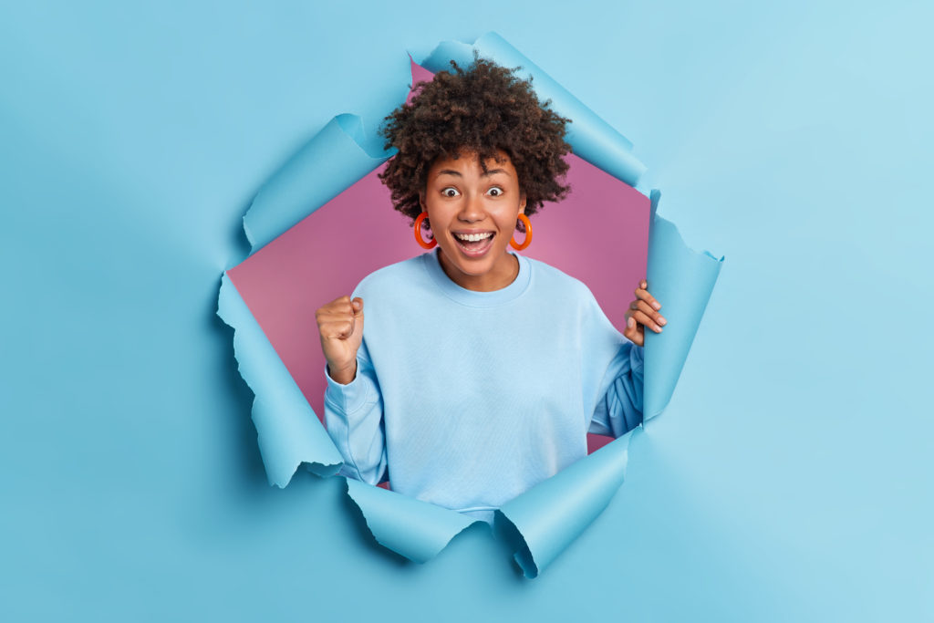 excited dark skinned young woman with curly hair clenches fists celebrates something awesome smiles broadly