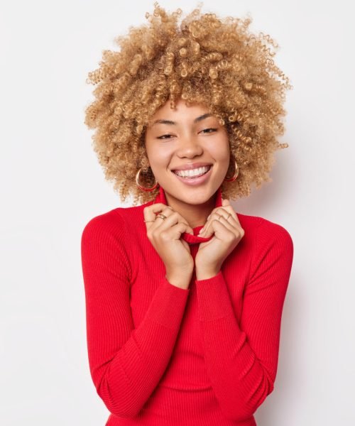 cheerful woman keeps hands on collar smiles pleasantly dressed in casual red turtleneck